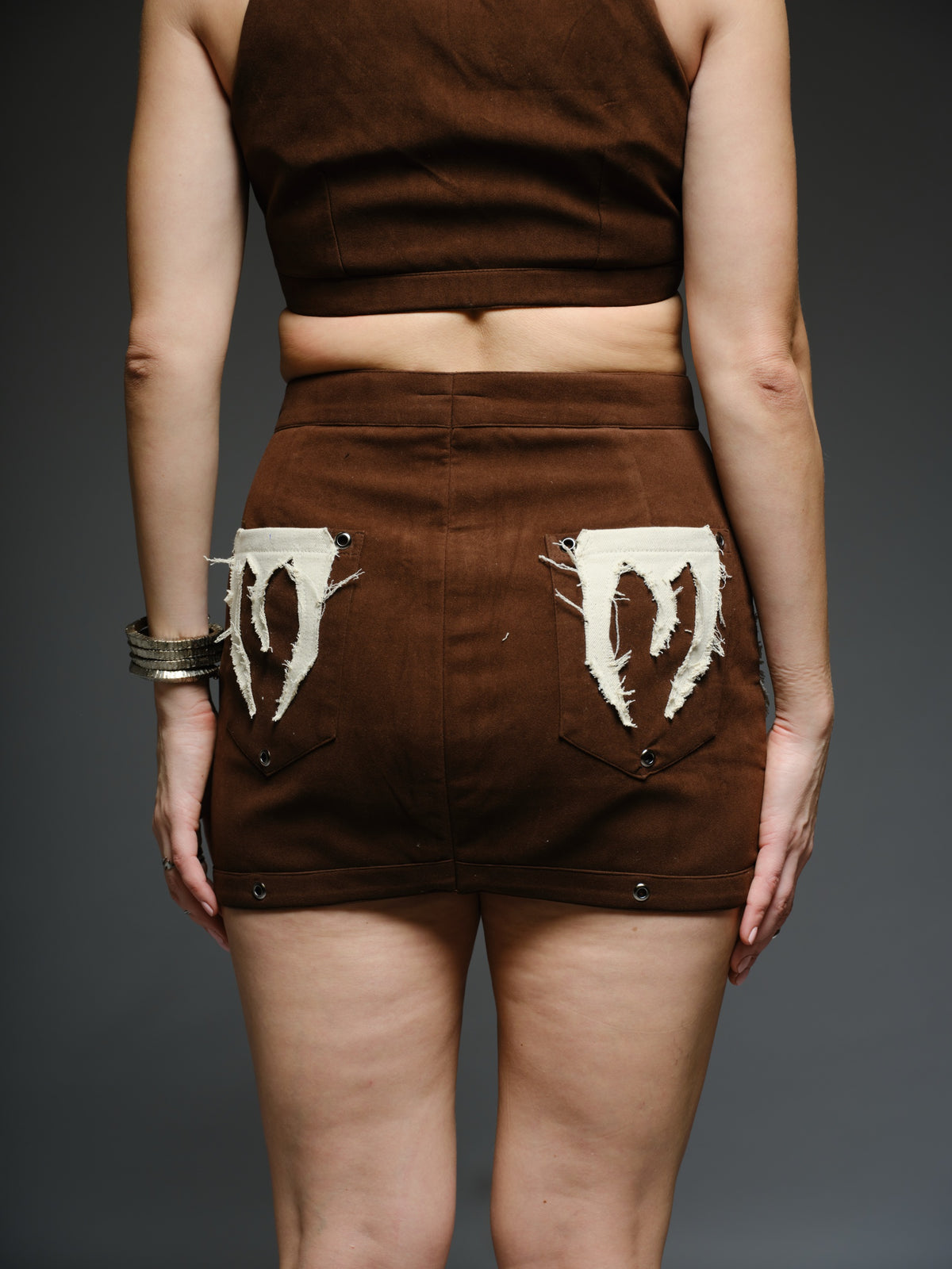 Brown suede collared crop top and mini skirt with angular white drill patches, frayed edges and silver-tone metal eyelets