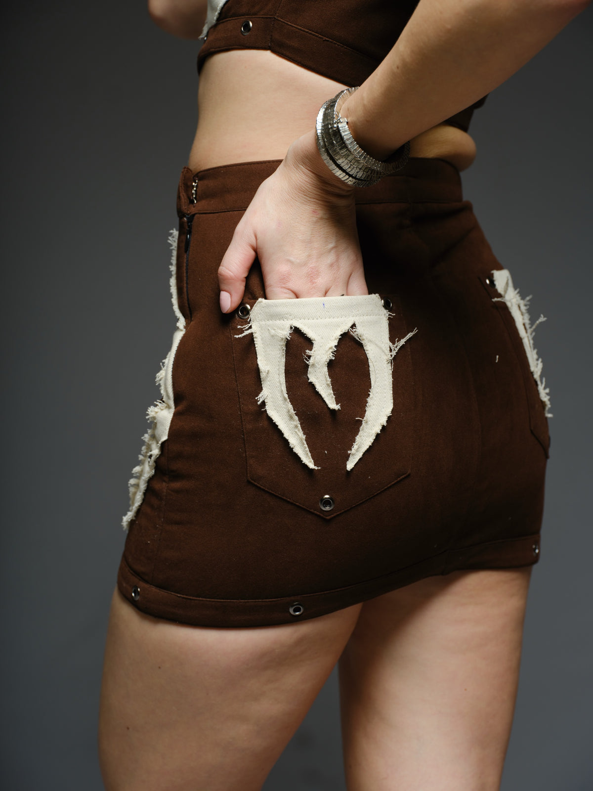 Brown suede collared crop top and mini skirt with angular white drill patches, frayed edges and silver-tone metal eyelets