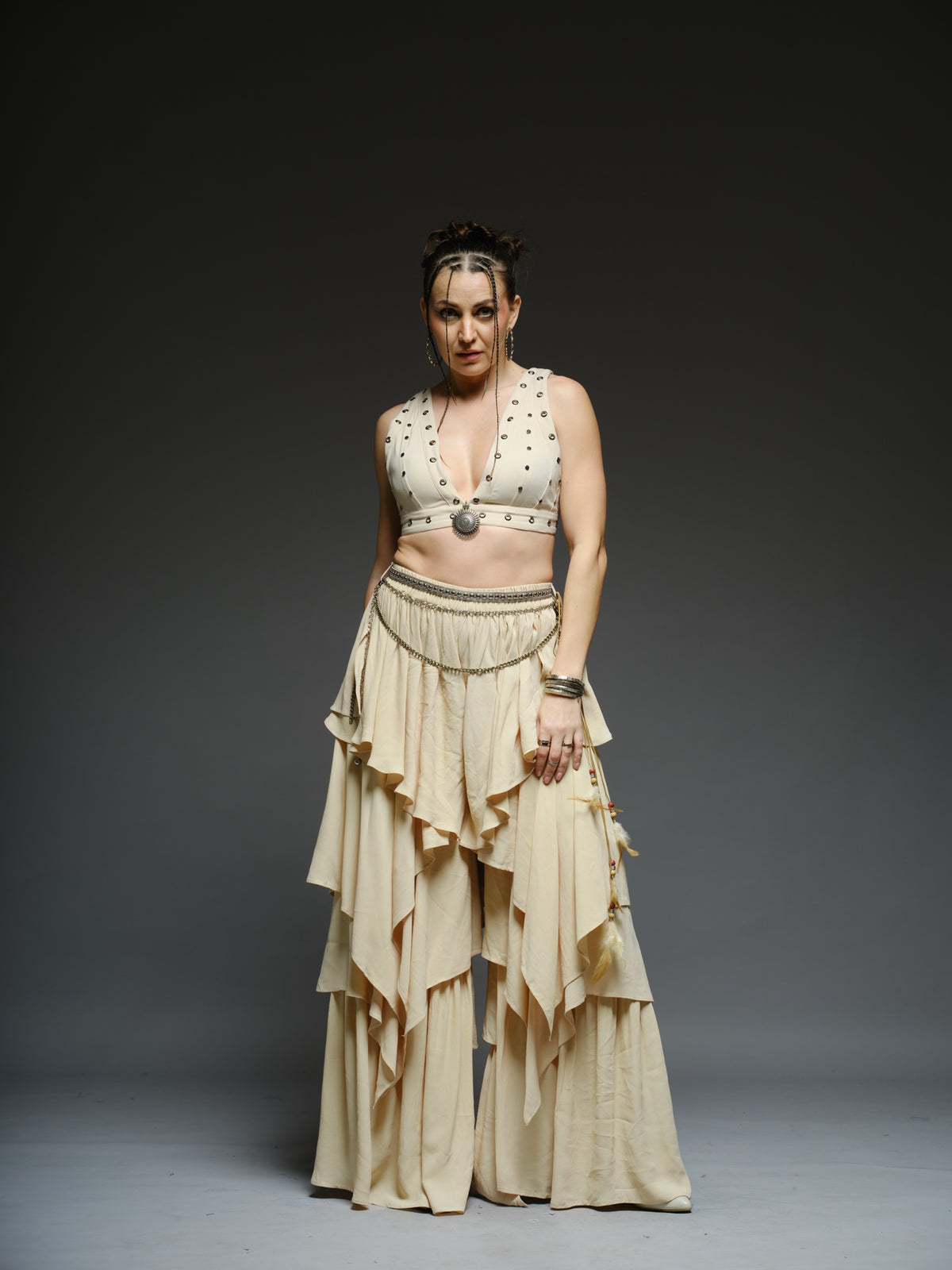 Beige studded V-neck bralette with a central brooch, paired with layered ruffle palazzo trousers with metallic belt and chain details