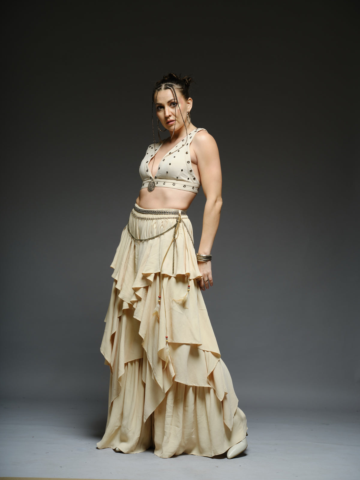 Beige studded V-neck bralette with a central brooch, paired with layered ruffle palazzo trousers with metallic belt and chain details