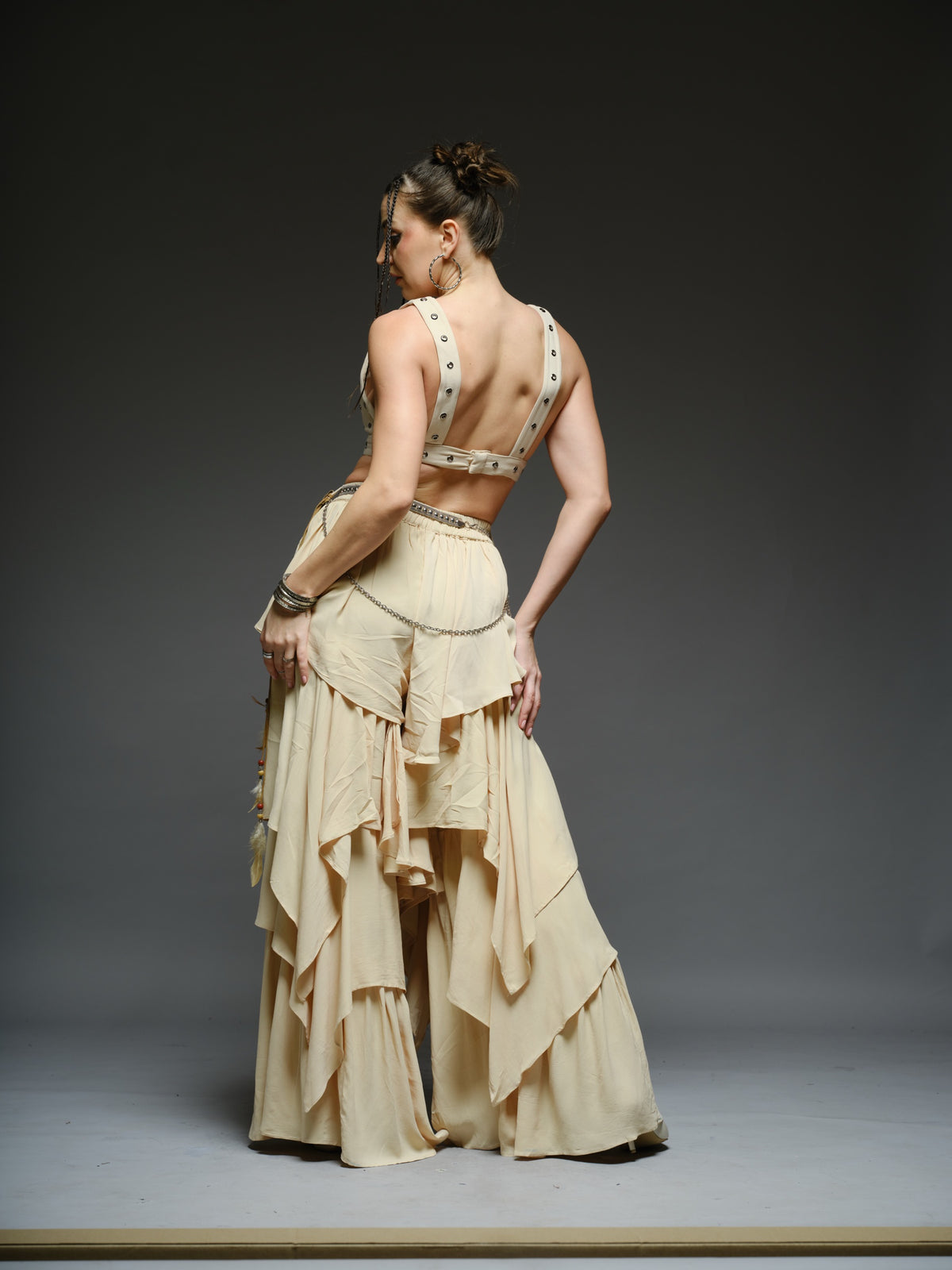 Beige studded V-neck bralette with a central brooch, paired with layered ruffle palazzo trousers with metallic belt and chain details