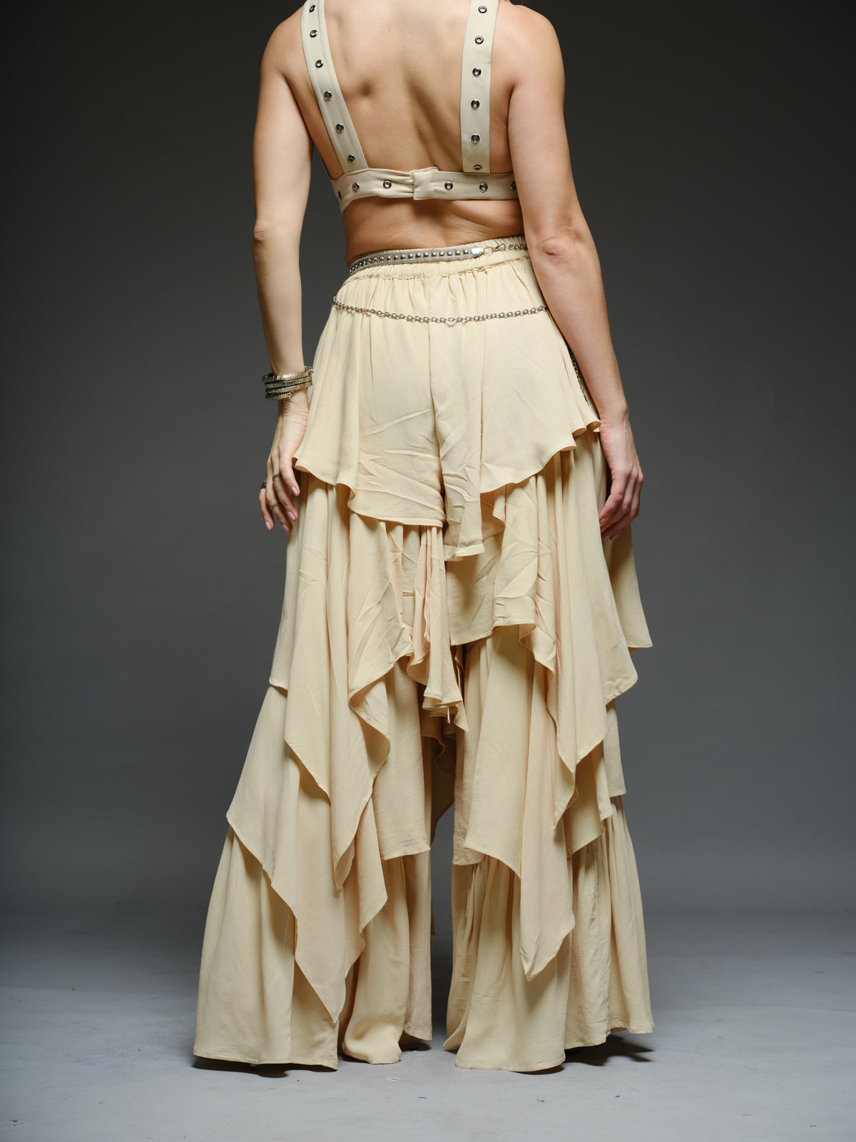Beige studded V-neck bralette with a central brooch, paired with layered ruffle palazzo trousers with metallic belt and chain details