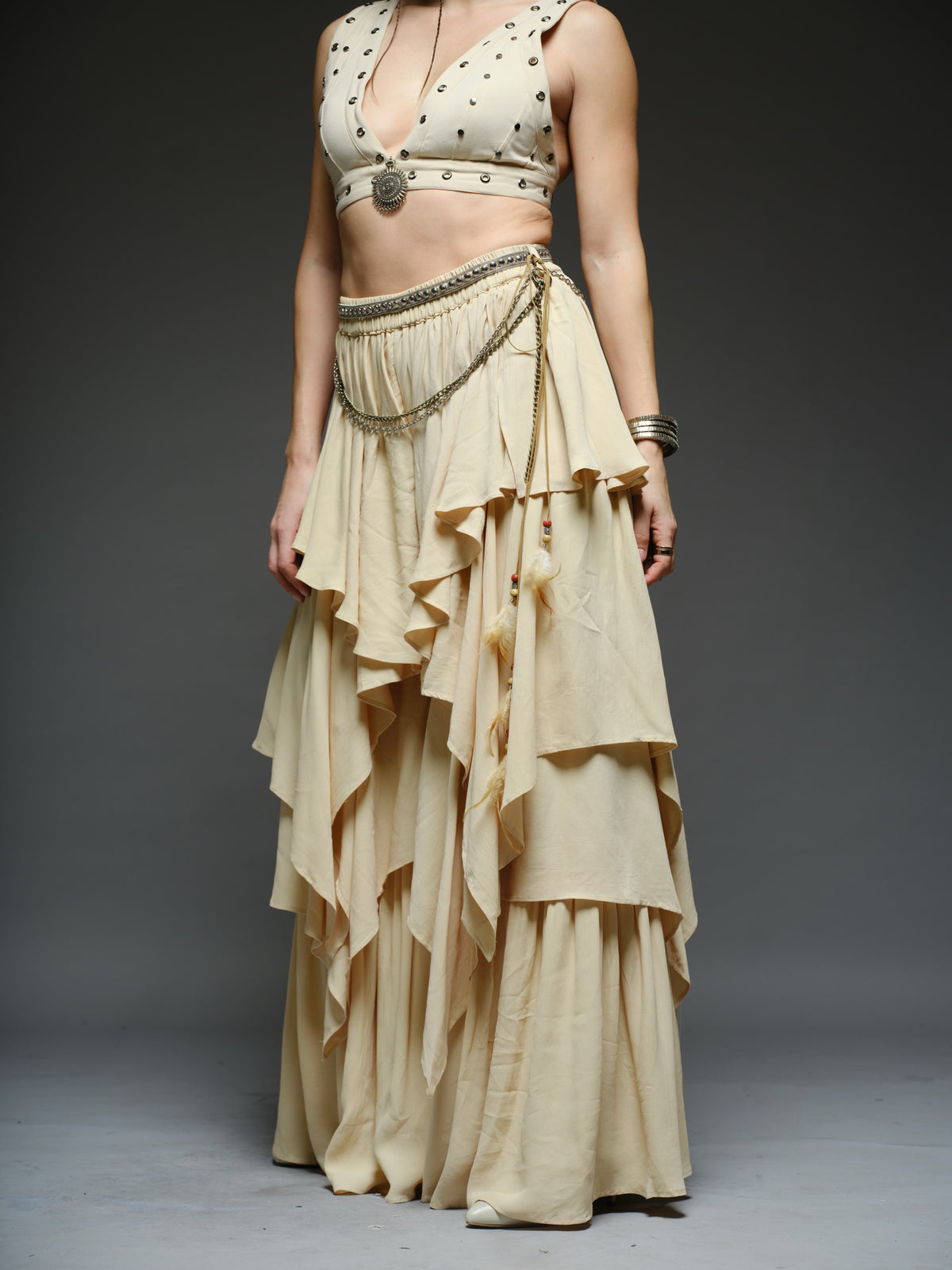 Beige studded V-neck bralette with a central brooch, paired with layered ruffle palazzo trousers with metallic belt and chain details