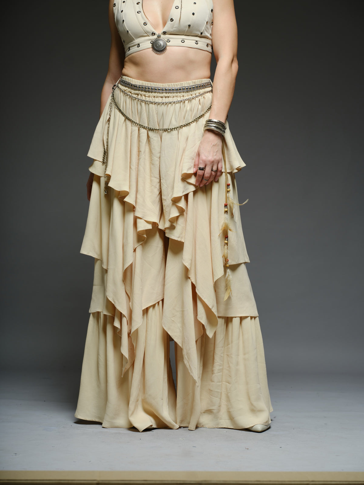 Beige studded V-neck bralette with a central brooch, paired with layered ruffle palazzo trousers with metallic belt and chain details