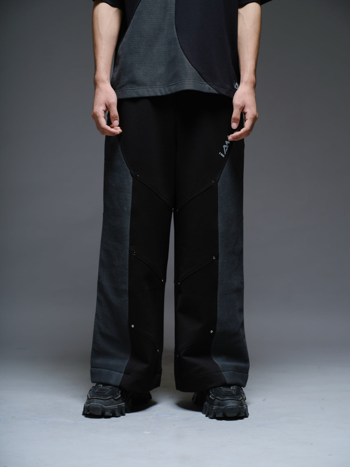 Loose black wide-leg trousers with grey net side panels and small silver-tone studs at the panel corners