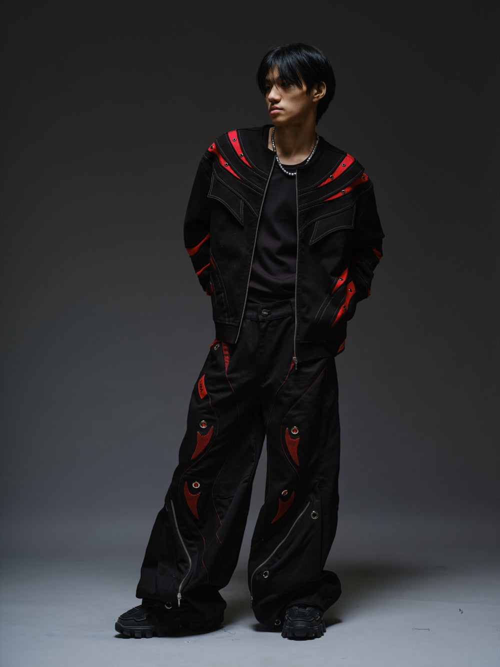 Black baggy denim pants with red patches beneath black net, oversized metal eyelets and large lower-leg zipper pockets