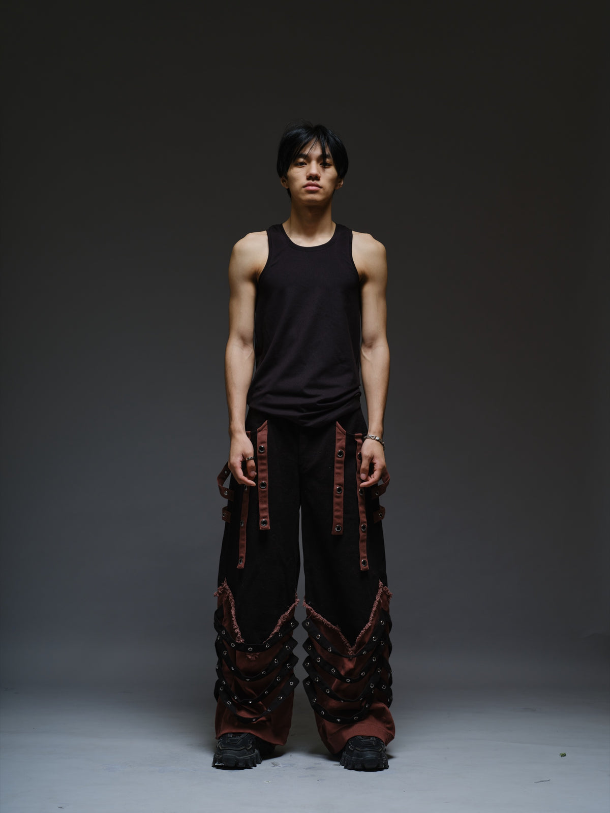 Black wide-leg baggy denim pants with brown hanging straps, layered lower-leg panels, metallic rivets and frayed brown trim