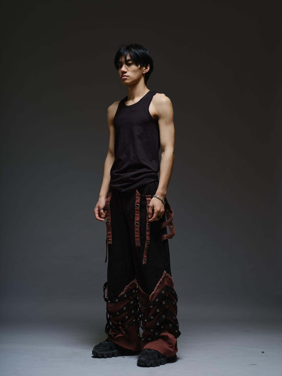 Black wide-leg baggy denim pants with brown hanging straps, layered lower-leg panels, metallic rivets and frayed brown trim