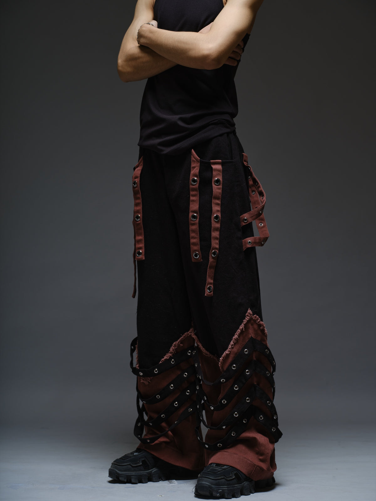 Black wide-leg baggy denim pants with brown hanging straps, layered lower-leg panels, metallic rivets and frayed brown trim