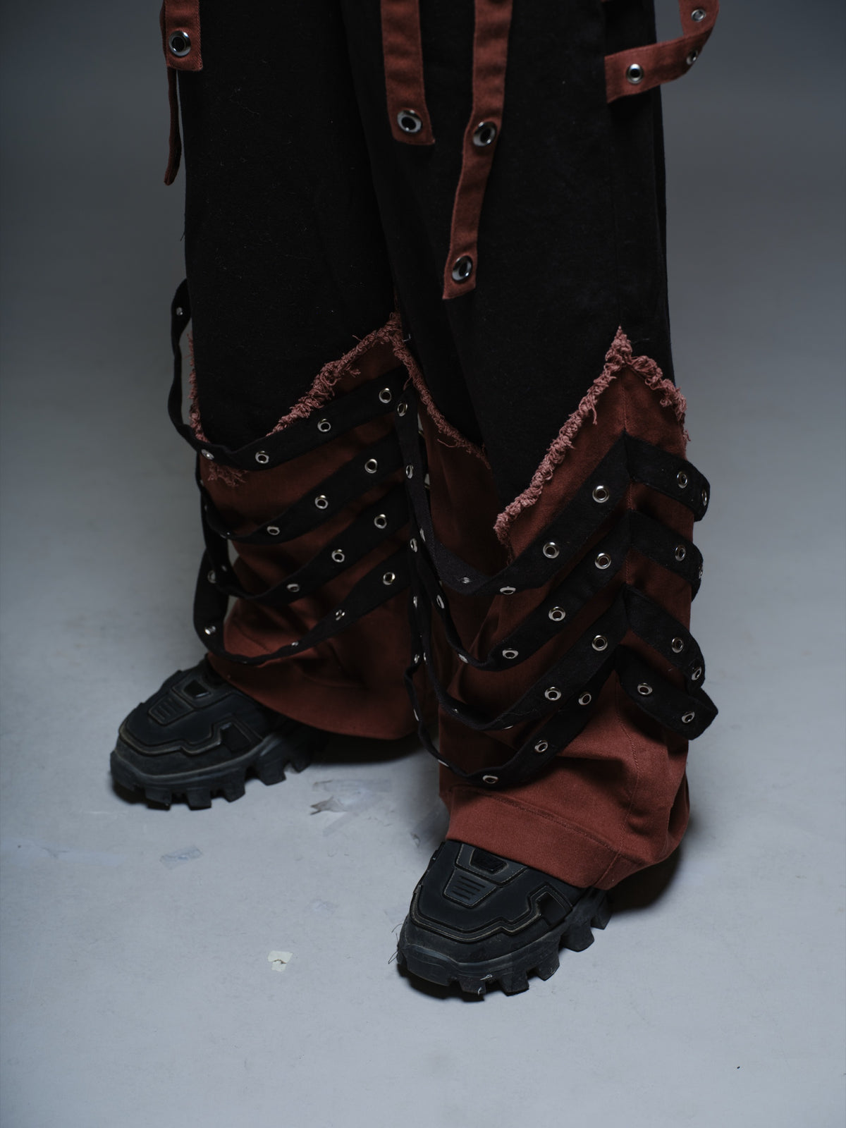 Black wide-leg baggy denim pants with brown hanging straps, layered lower-leg panels, metallic rivets and frayed brown trim