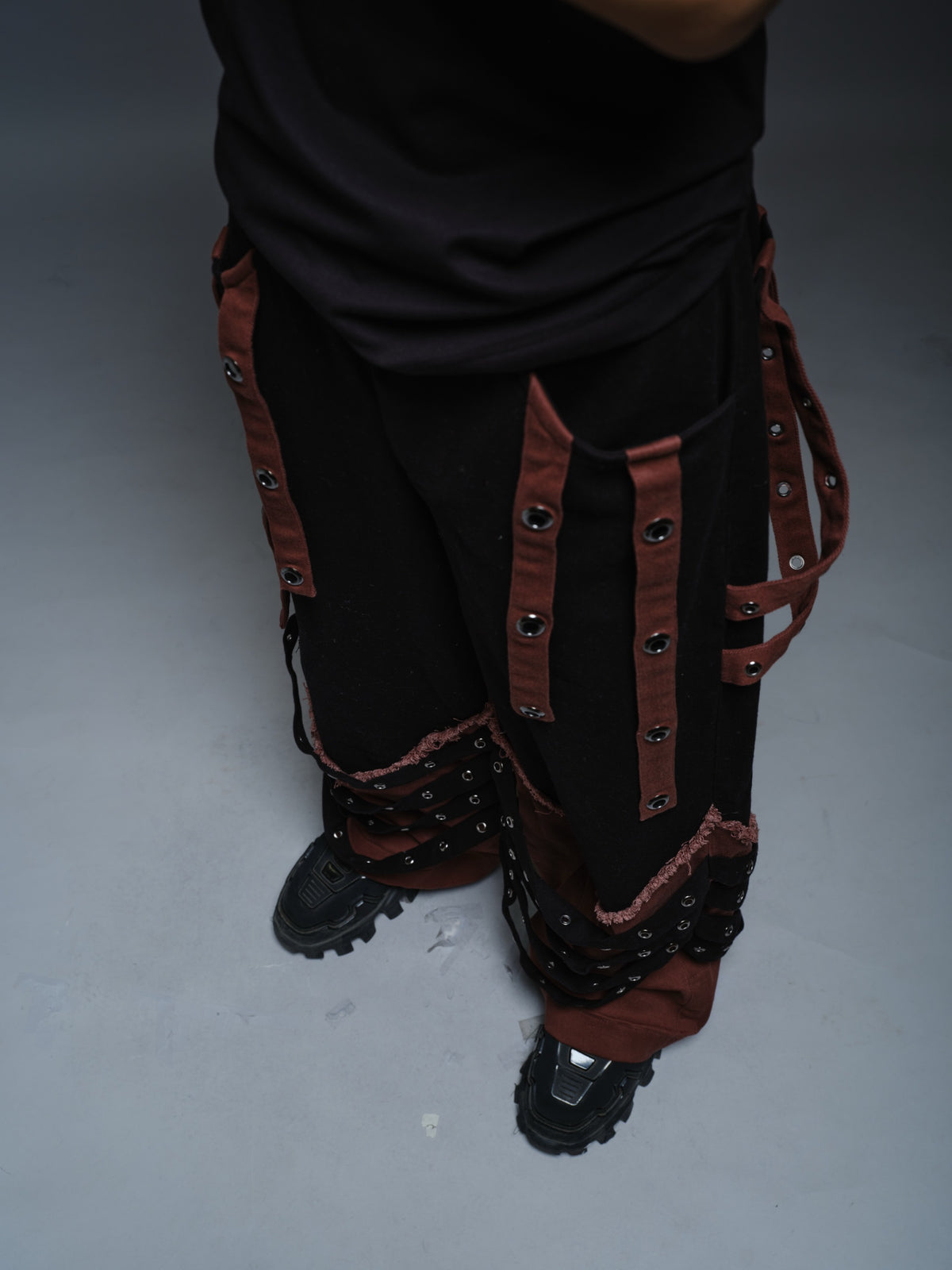 Black wide-leg baggy denim pants with brown hanging straps, layered lower-leg panels, metallic rivets and frayed brown trim
