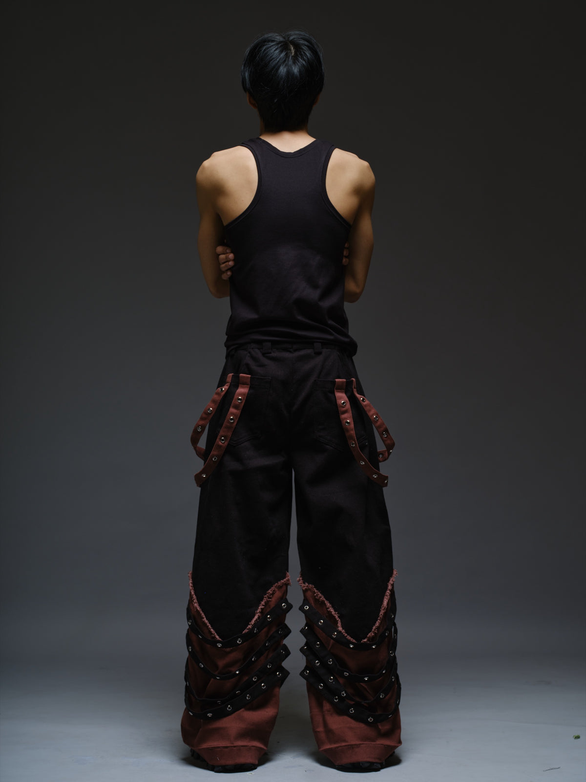 Black wide-leg baggy denim pants with brown hanging straps, layered lower-leg panels, metallic rivets and frayed brown trim