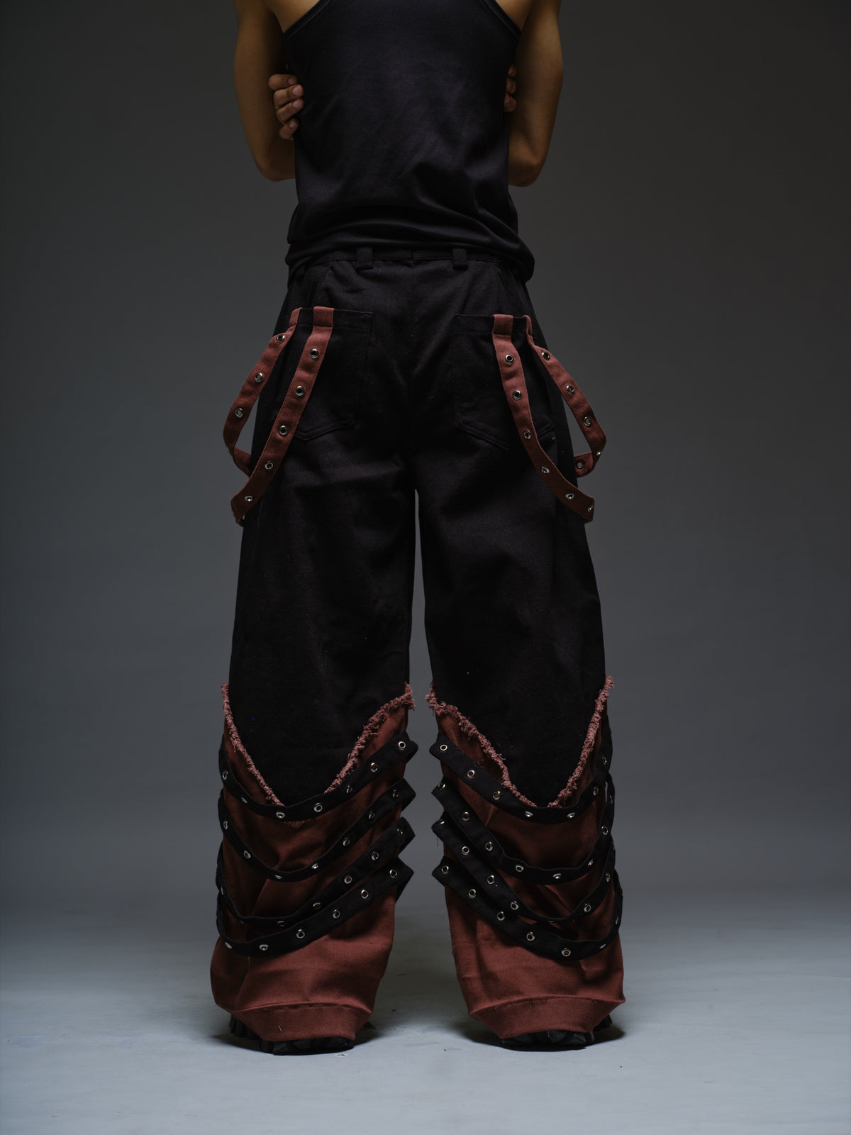 Black wide-leg baggy denim pants with brown hanging straps, layered lower-leg panels, metallic rivets and frayed brown trim