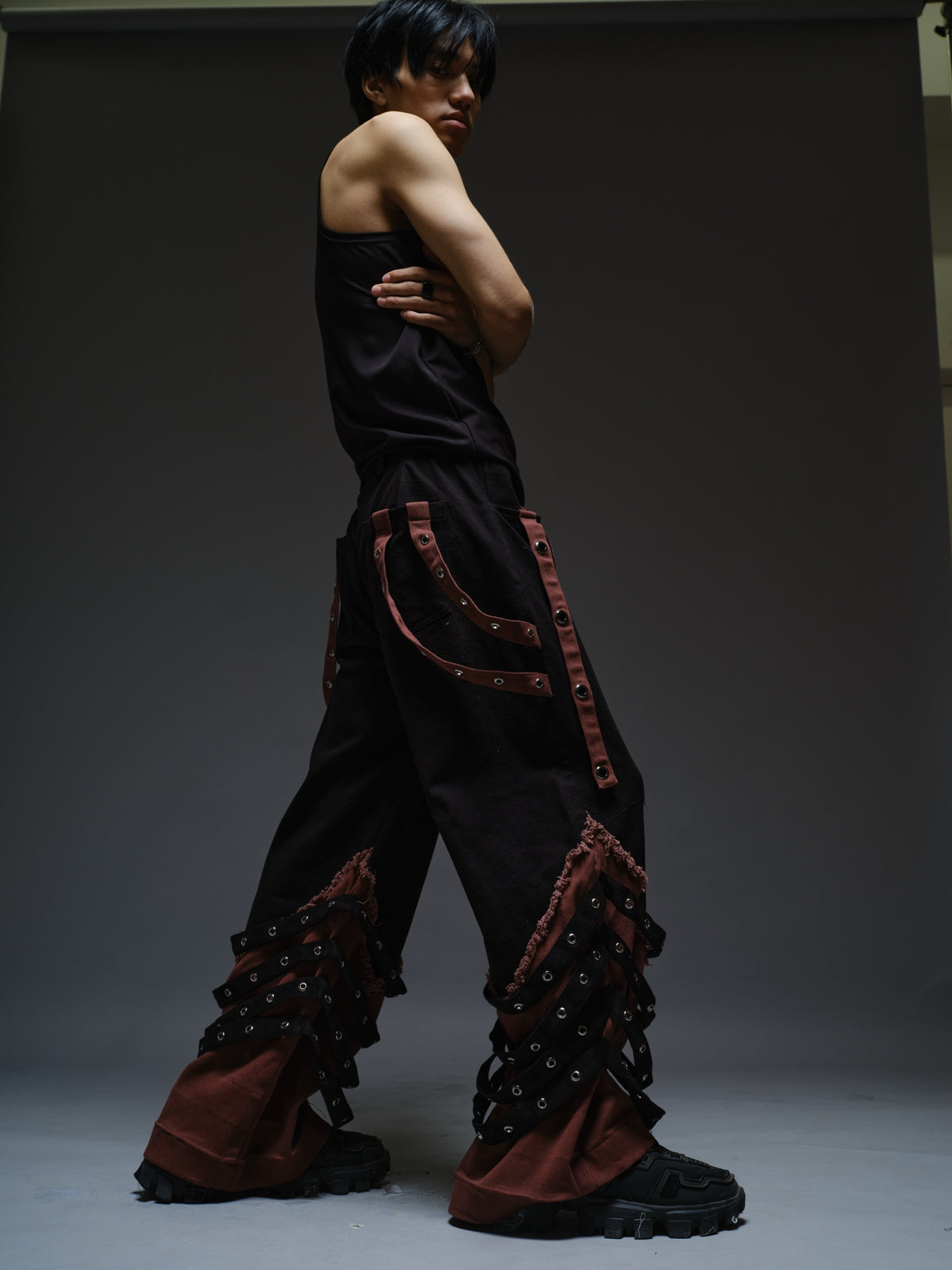 Black wide-leg baggy denim pants with brown hanging straps, layered lower-leg panels, metallic rivets and frayed brown trim